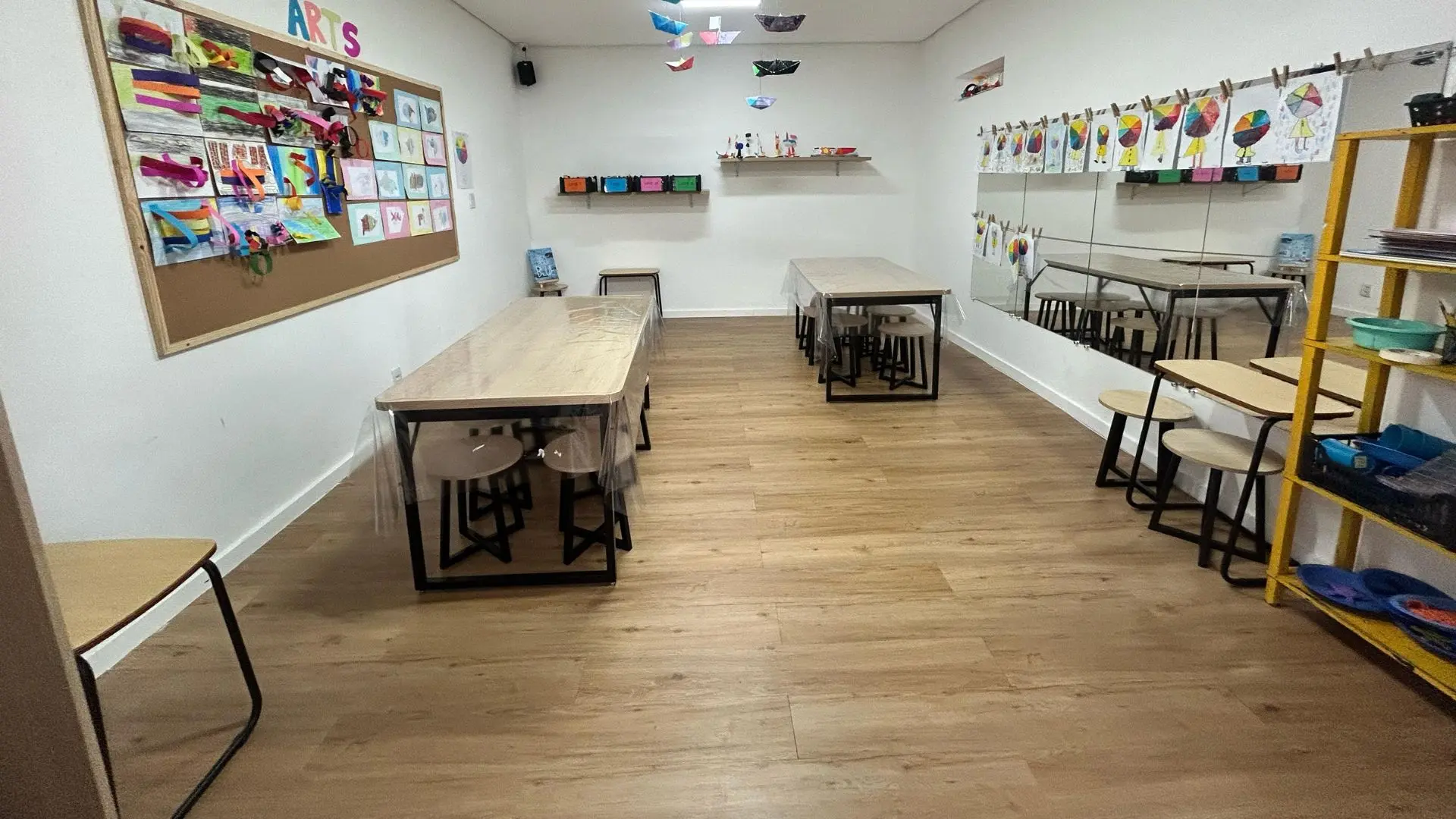Sala de artes da Trails School, com mural de trabalhos dos alunos, mesas, banquetas e estante de materiais